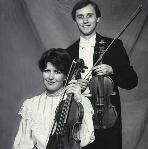 A black and white photo of a young man wearing a tuxedo, standing with his violin. In front of this is a young woman, wearing a white frilly dress holding a viola