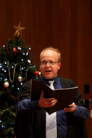 Man wearing glasses and holding a black folder while singing in front of a Christmas tree