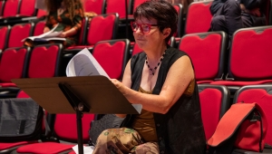 Woman with short brown hair and red glasses sitting on a red chair reading music from a music stand