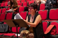 Woman with short brown hair and red glasses sitting on a red chair reading music from a music stand