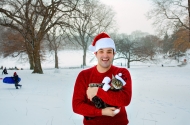 Young man standing in the snow wearing a red sweater and a santa hat, holding a tabby cat in his arms. The cat is wearing a santa outfit.
