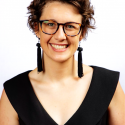Young woman with short brown hair and glasses weaing long dangly earrings and a black dress
